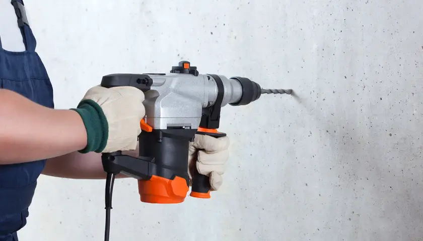 Une personne en salopette bleue, portant des gants, utilise un perforateur pour percer un mur en béton.