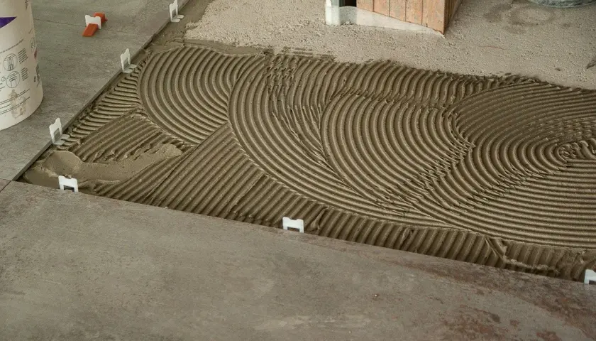 La chape est prête à recevoir les carreaux, avec un mortier étalé en ondulations parfaites. Des croisillons blancs maintiennent l'espacement.