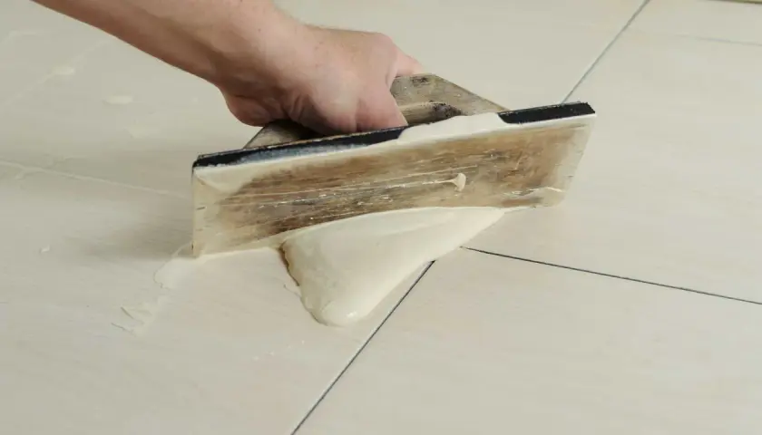 Main tenant utilise une spatule en bois pour faire des joints de carrelage, étalant le mortier sur les carreaux clairs.