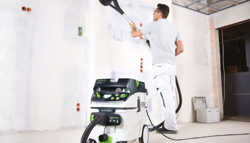 Un ouvrier utilise un aspirateur pour girafe afin de poncer un mur.