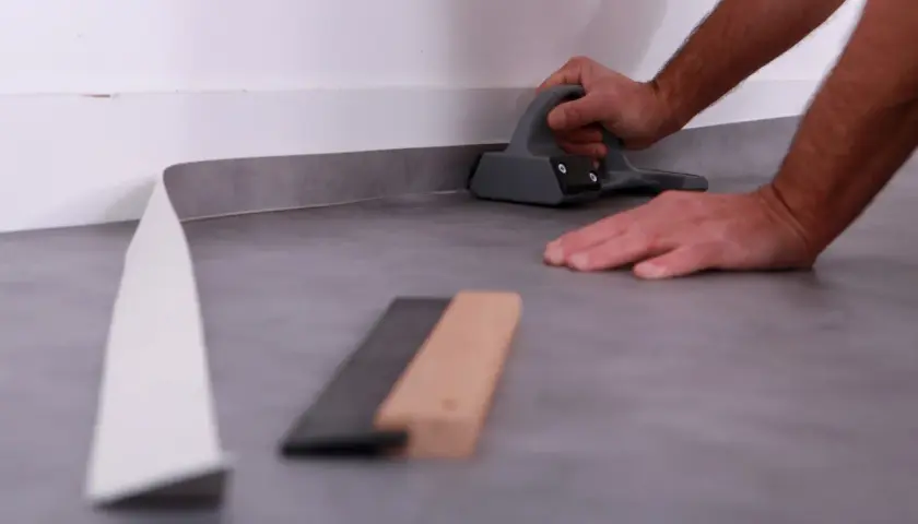 Main tenant une raclette pour bien comment poser du lino. Des outils sont posés au sol.