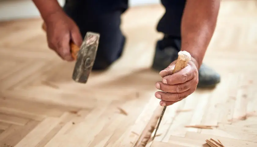 Un artisan utilise un ciseau et un marteau pour changer une lame de parquet flottant endommagée.