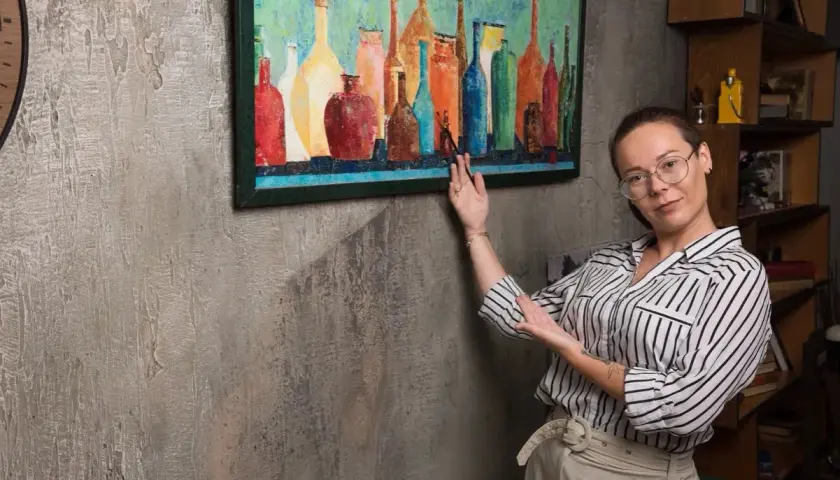 Une femme montre comment accrocher un cadre coloré de bouteilles sur un mur texturé.