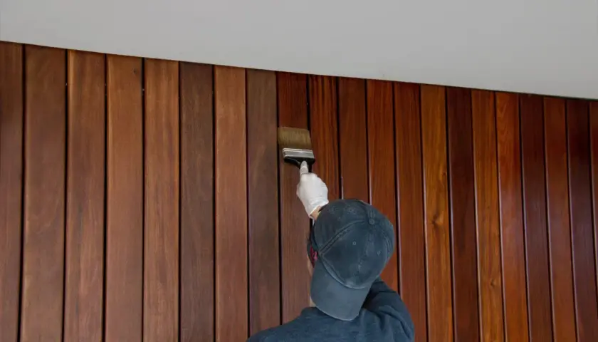 Une personne porte un gant blanc et un casquette pour peindre du lambris en bois avec un pinceau.