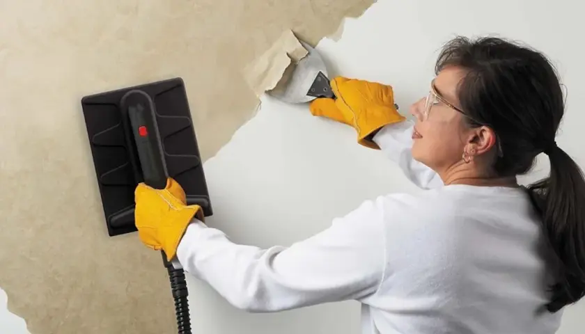 Une femme, portant des gants jaunes, utilise un appareil pour décoller du papier peint ancien. Elle gratte la surface avec un outil.