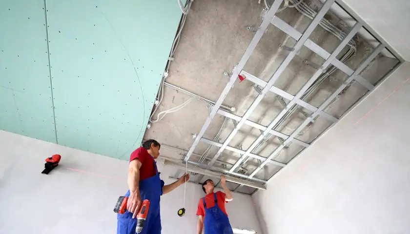 Deux ouvriers installent une structure métallique pour un schema montage placo plafond. Des fils électriques sont déjà en place.