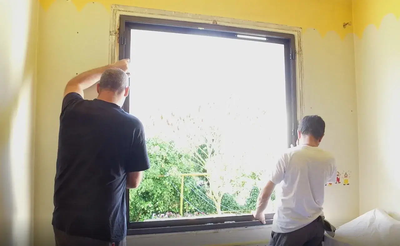 Deux hommes installent une nouvelle fenêtre. La lumière du jour filtre à travers le vitrage, révélant un jardin verdoyant. La pièce est encore en travaux, mais bientôt, cette fenêtre dormante s'ouvrira sur le monde.