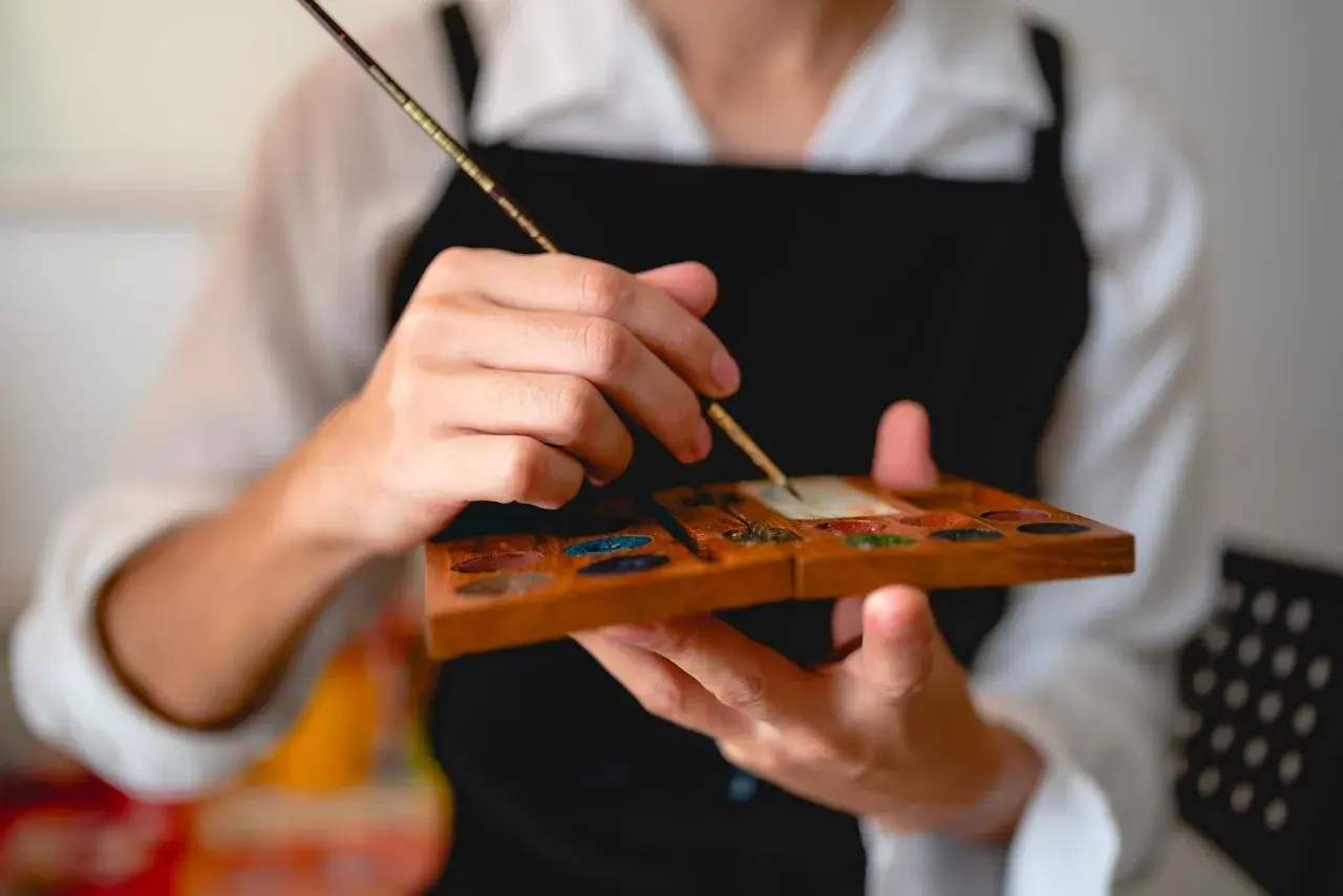 Main tenant une palette en bois remplie de couleurs, un artiste utilise un pinceau, un des outils pour peindre, pour mélanger des pigments.