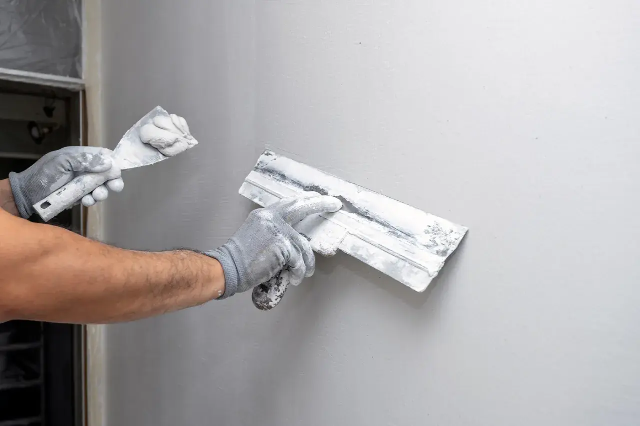 Mains gantées appliquant de l'enduit sur un mur gris, étape clé de la construction murale.