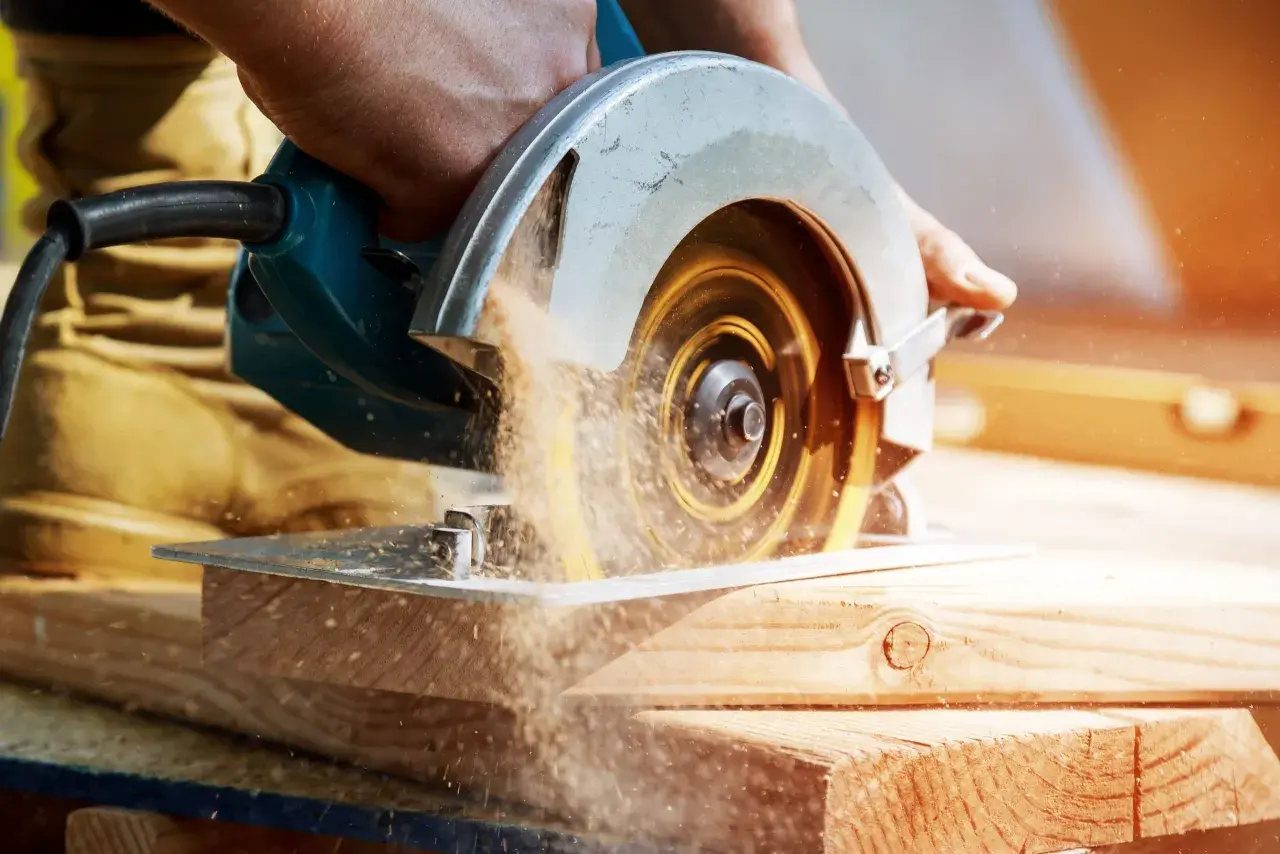 Main d'un bricoleur utilisant une scie circulaire pour couper du bois. La poussière de bois vole autour de la lame. Apprenez comment affûter une lame de scie circulaire pour des coupes nettes.