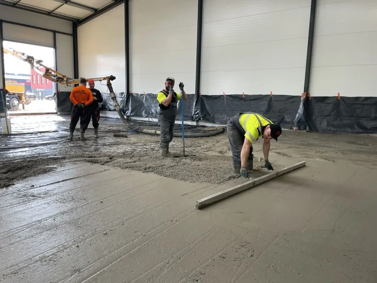 Des ouvriers coulent et lissent du béton frais sur un sol, avec une pompe à béton en arrière-plan.