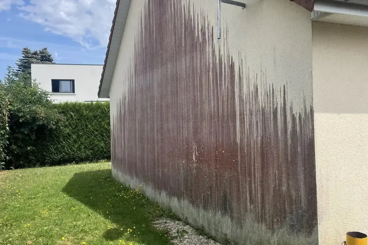 Le mur extérieur d'une maison présente une importante moisissure rouge, formant des traînées sombres sur le crépi clair.