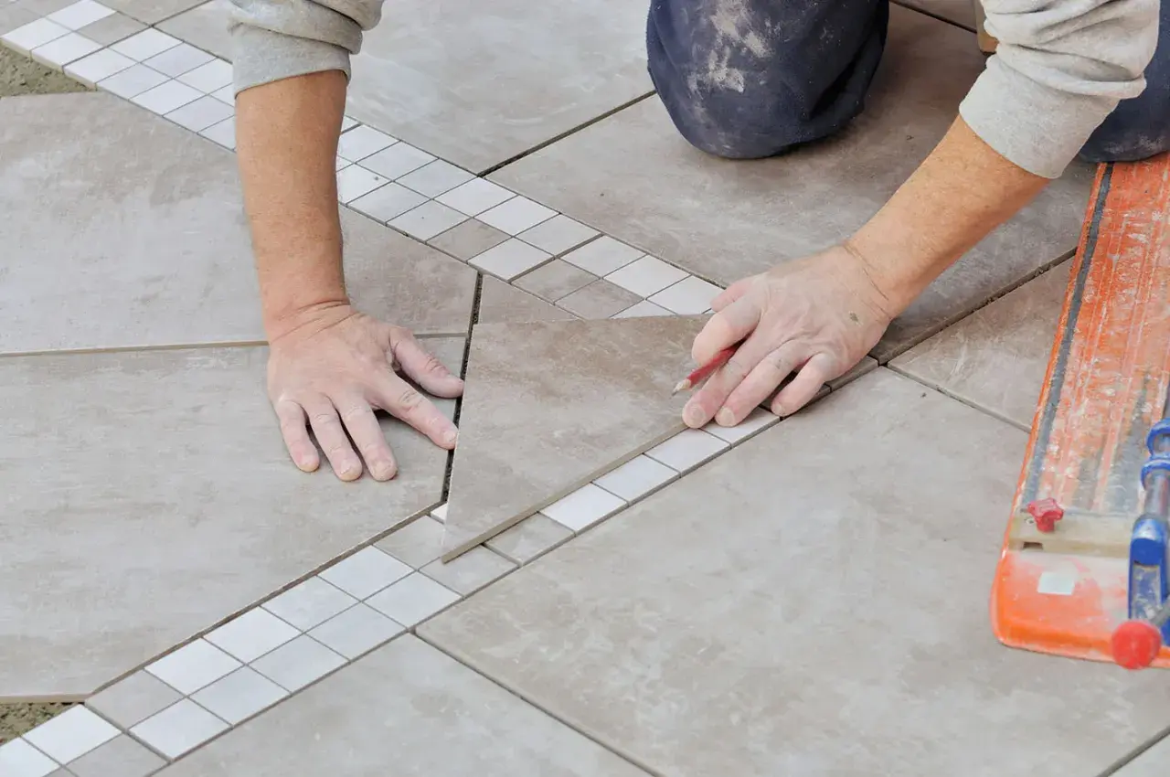 Mains expertes en pose carrelage, ajustant une pièce avec précision. Un coupe-carreau orange est visible.