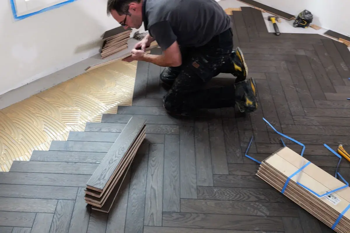 Pose de parquet en pose bâton rompu. Un artisan applique de la colle sur le sol avant de poser les lames de bois.