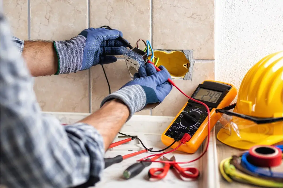 Un électricien vérifie le **fonctionnement d'une gâche électrique** avec un multimètre. Outils et casque de sécurité à proximité.