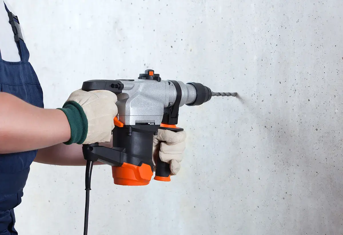 Une personne en salopette bleue, portant des gants, utilise un perforateur pour percer un mur en béton.