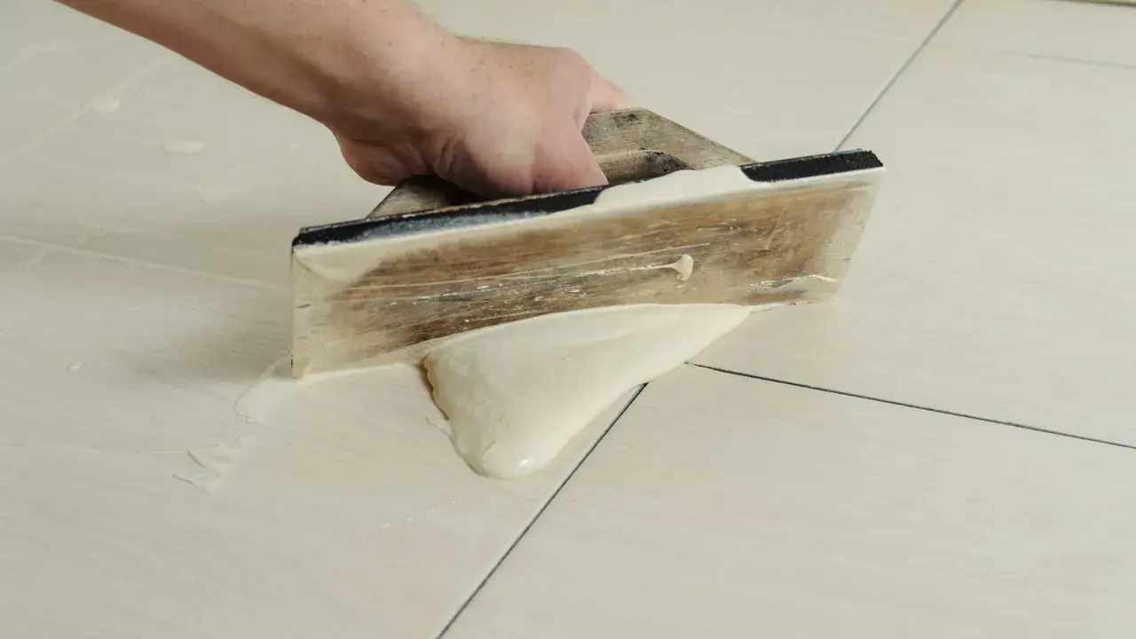 Main tenant utilise une spatule en bois pour faire des joints de carrelage, étalant le mortier sur les carreaux clairs.
