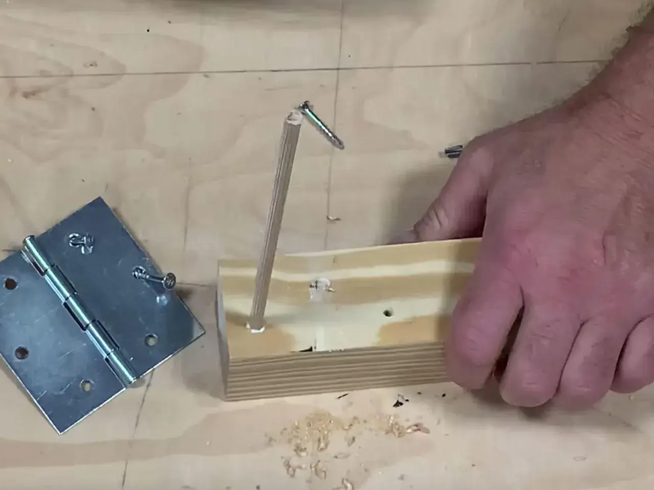 Un bricoleur teste un montage : une vis qui tourne dans le vide, tenue par un bout de bois, à côté d'une charnière.