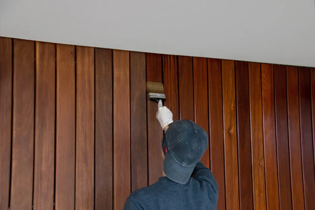 Une personne porte un gant blanc et un casquette pour peindre du lambris en bois avec un pinceau.