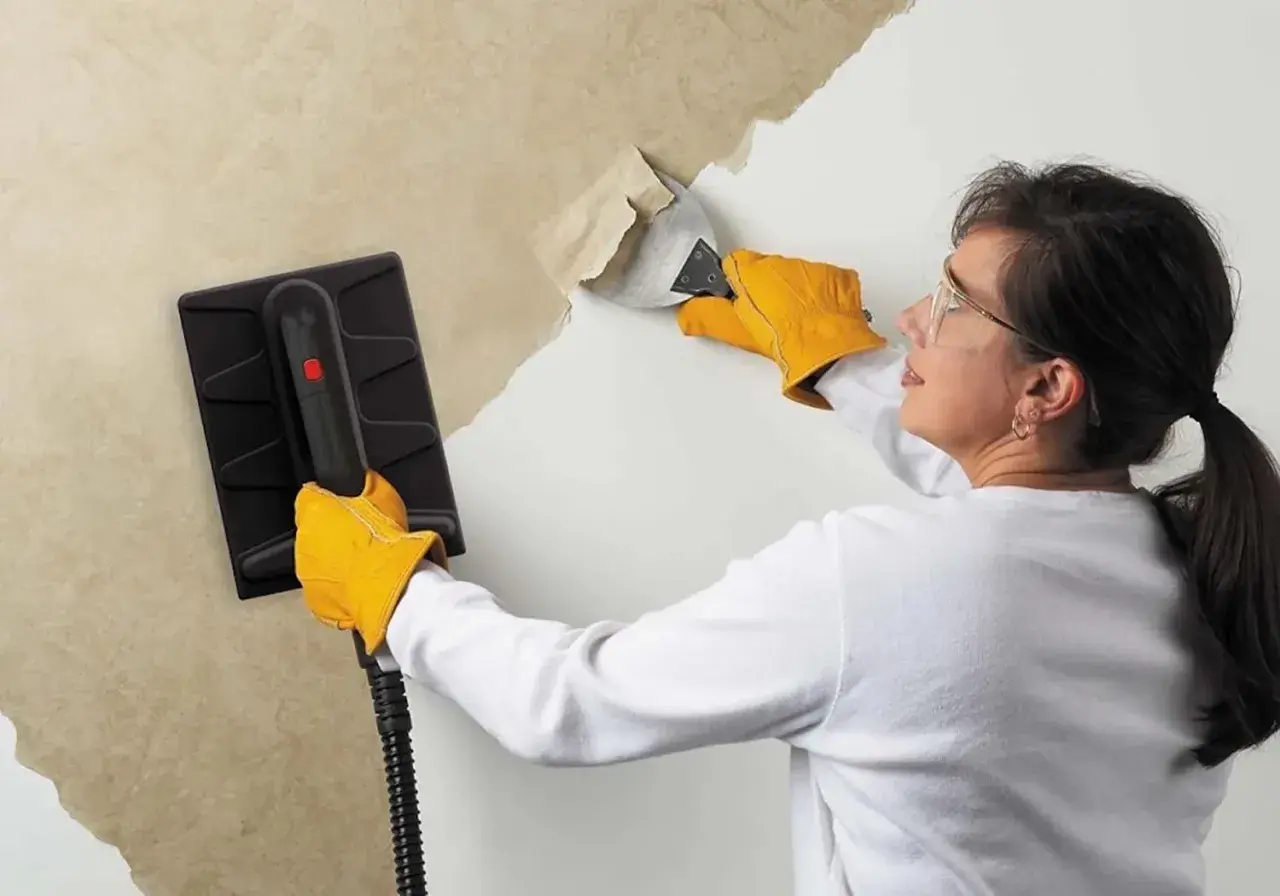 Une femme, portant des gants jaunes, utilise un appareil pour décoller du papier peint ancien. Elle gratte la surface avec un outil.