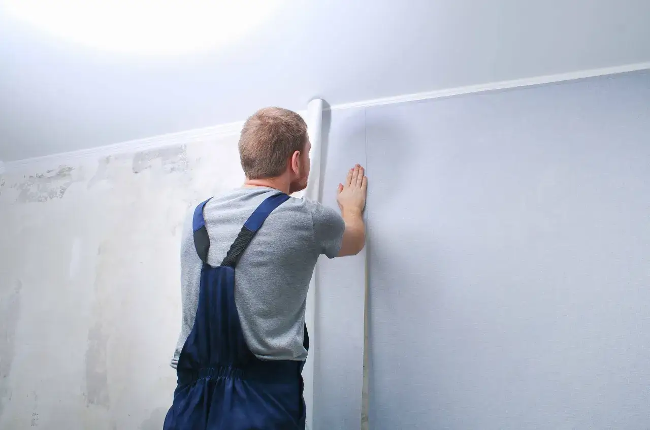 Un homme en salopette bleue est en train de d&eacute;coller du papier peint sur un mur. La partie gauche du mur est d&eacute;j&agrave; d&eacute;nud&eacute;e, r&eacute;v&eacute;lant l'ancienne peinture.