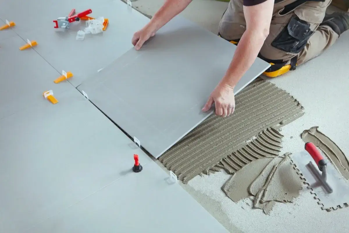 Pose d'une nouvelle dalle de carrelage gris sur du mortier frais, avec des cales et des outils de carreleur visibles.