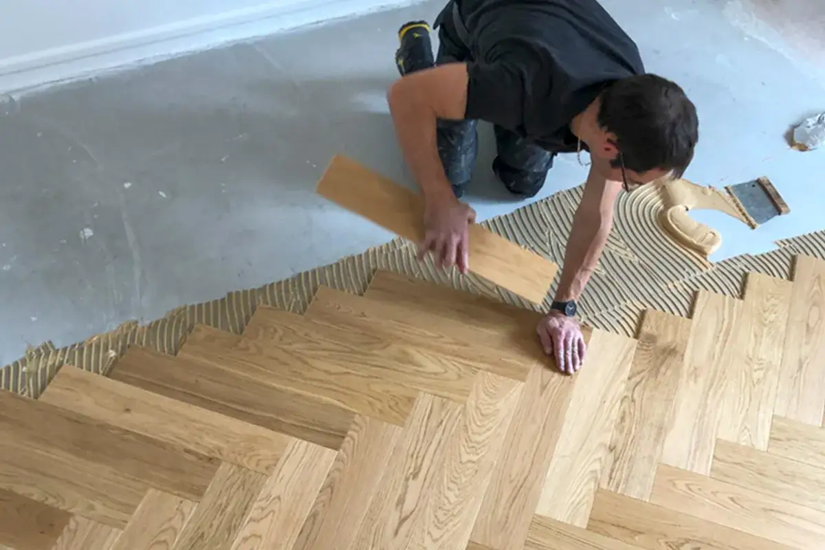 Pose baton rompu : un artisan pose un parquet en bois, créant un motif en chevrons sur un sol préparé avec de la colle.