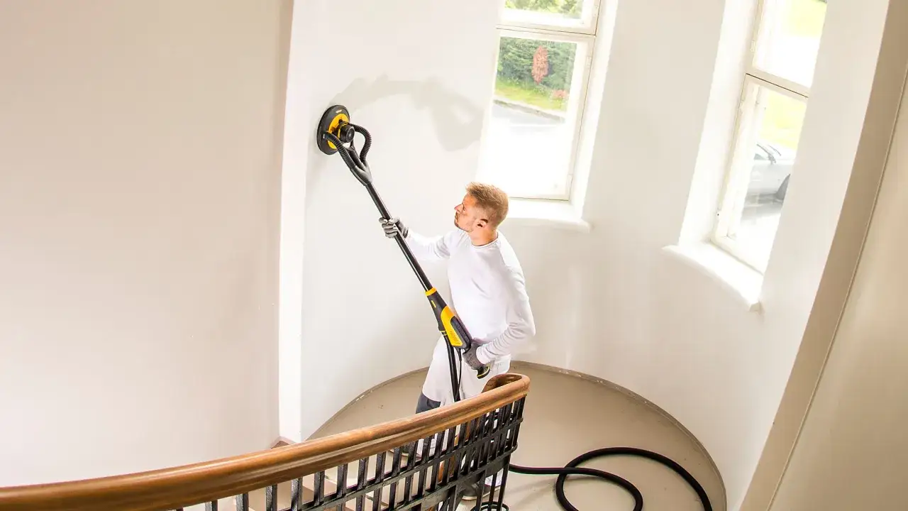 Un homme utilise une ponceuse pour enlever du crépi intérieur sur un mur blanc, près d'un escalier en colimaçon et de fenêtres.