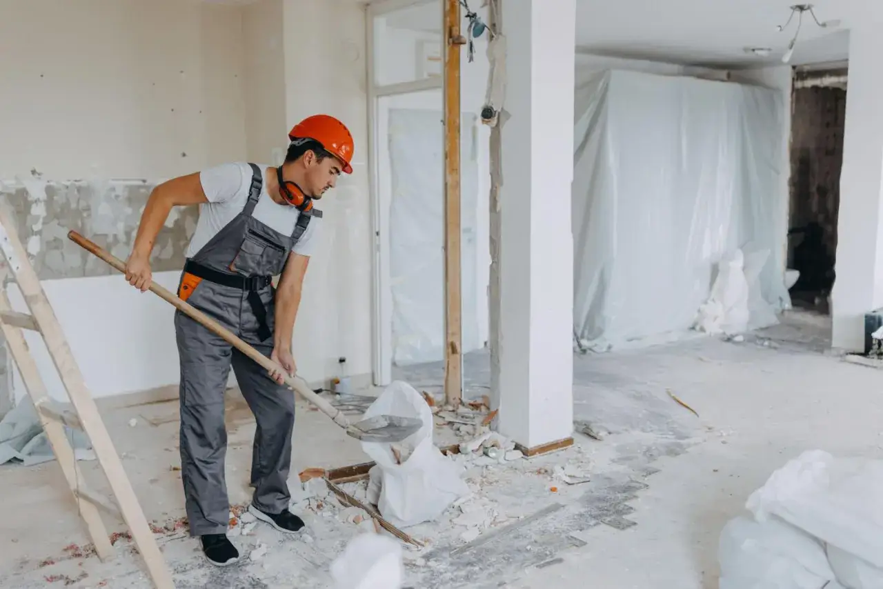 Un ouvrier en casque orange pelle des débris. La poussière de plâtre est omniprésente, un danger à ne pas négliger lors de ces travaux.