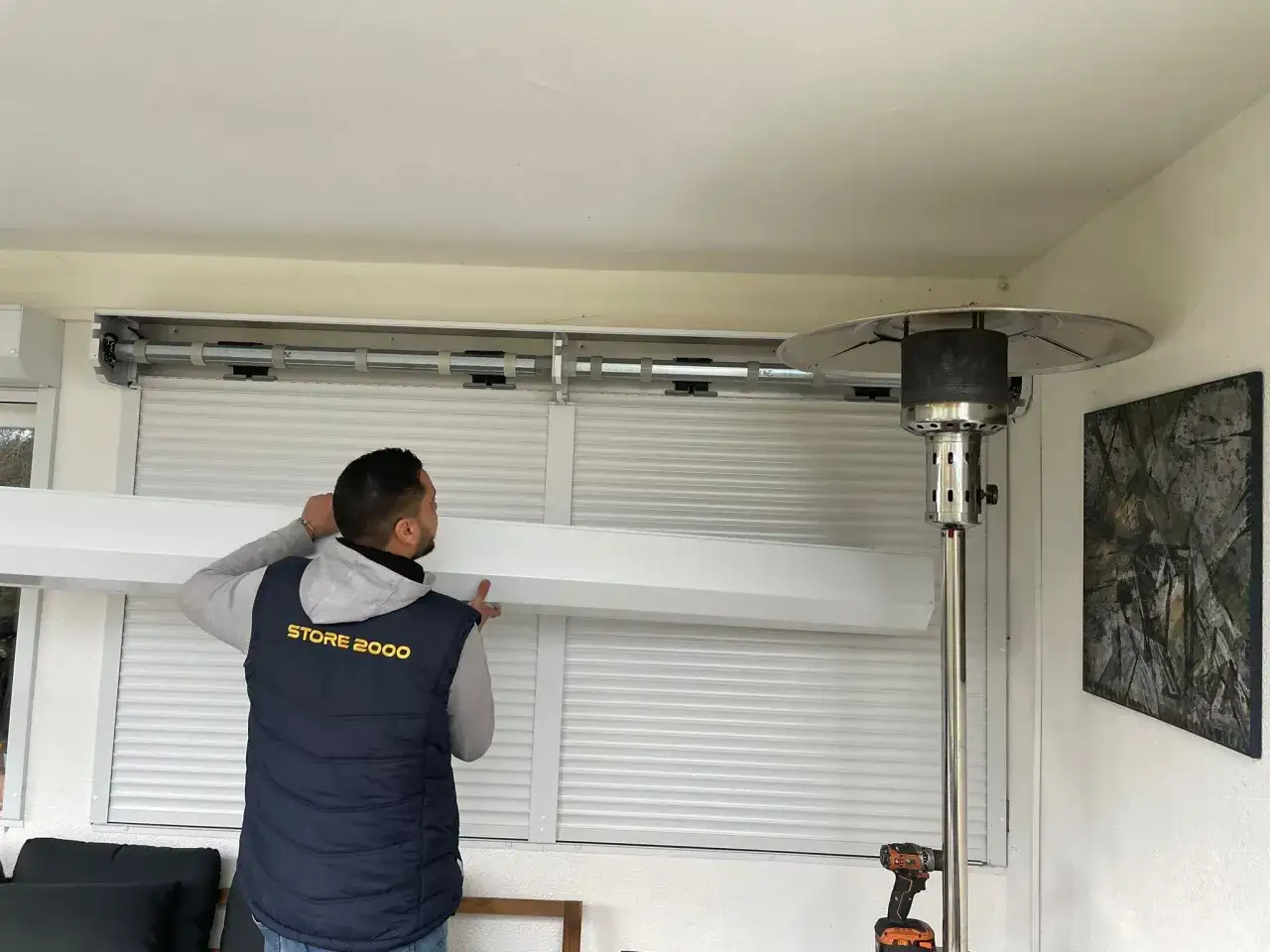 Un technicien installe un système pour motoriser des volets roulants. Un chauffage d'extérieur est visible à droite.
