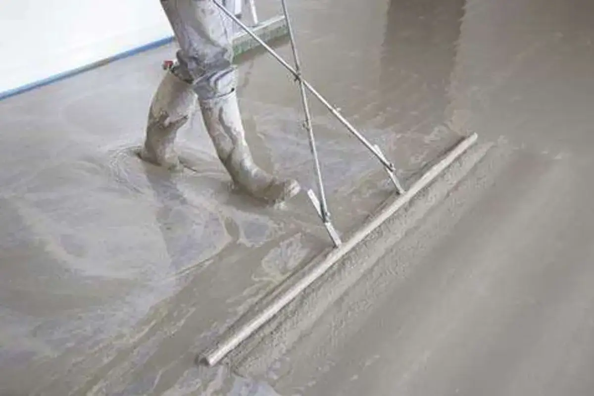 Un ouvrier en bottes travaille sur une chape fraîchement coulée, utilisant une règle de maçon pour lisser le béton.
