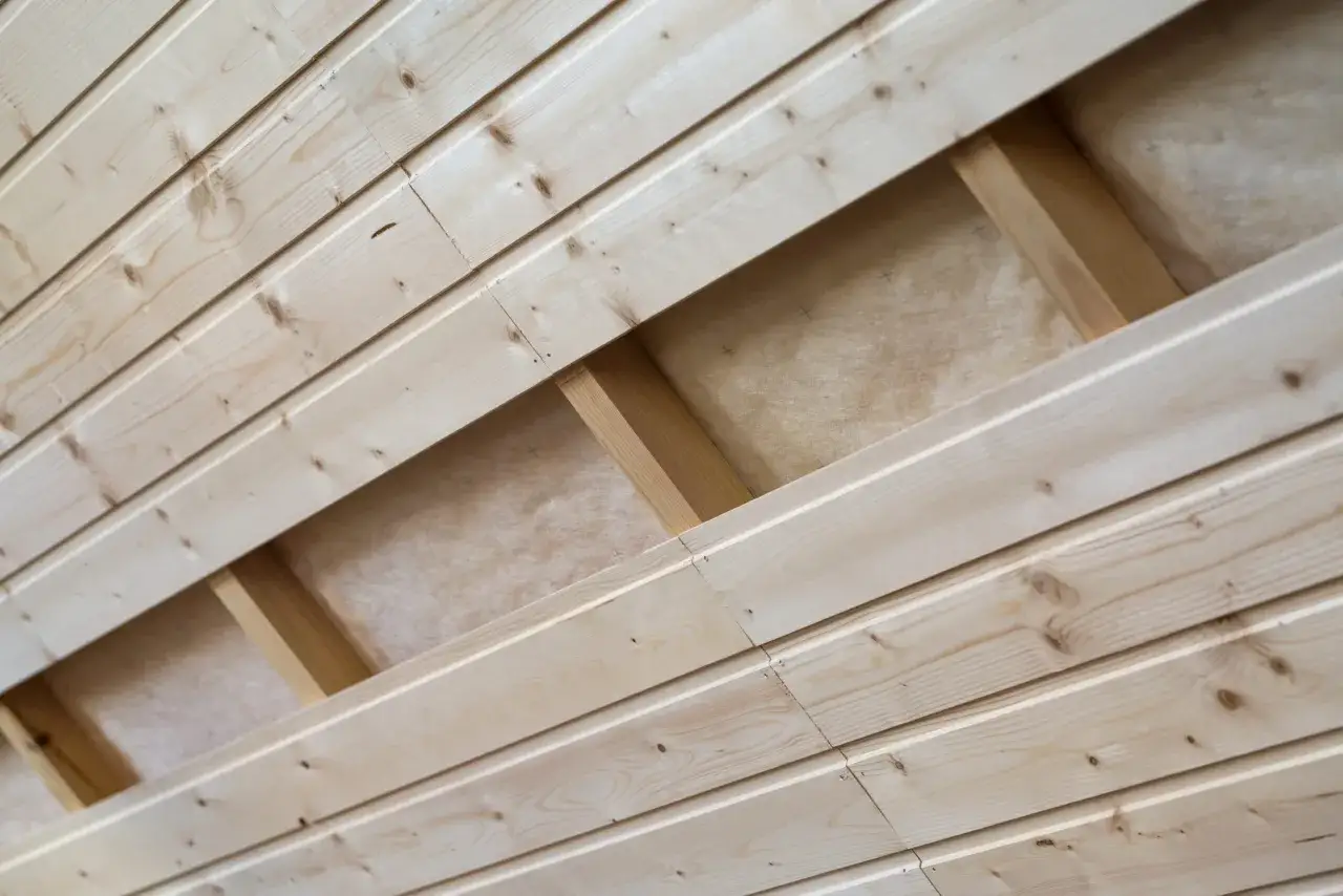 Pose lambris en bois clair, montrant la structure sous-jacente et l'isolant. Les planches sont assemblées en diagonale, créant un motif naturel.