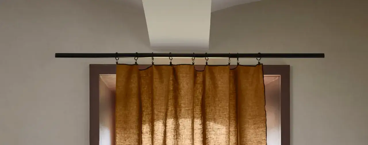 Une tringle &agrave; rideaux noire est fix&eacute;e au mur au-dessus d'une fen&ecirc;tre. Des anneaux noirs sont accroch&eacute;s &agrave; la tringle, supportant un rideau en lin brun.