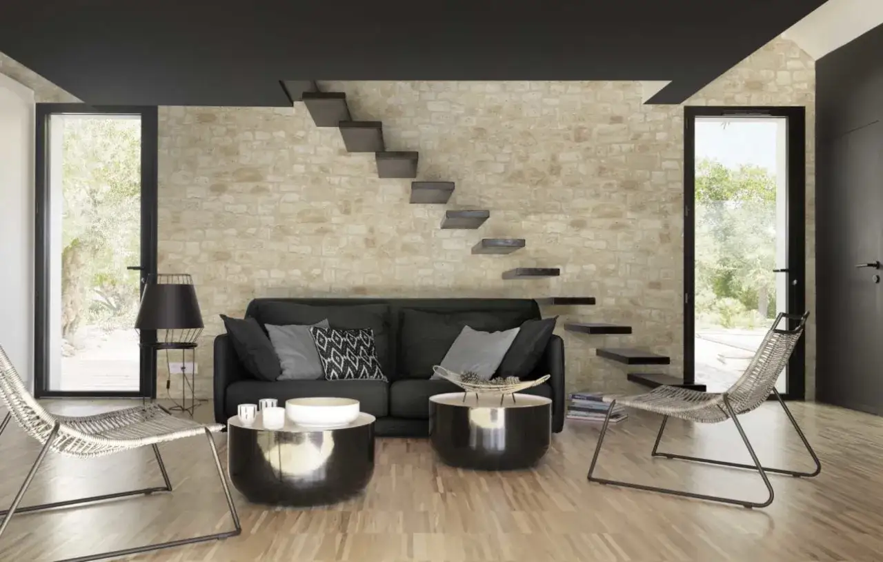 Salon moderne avec mur en pierre de parement intérieur, escalier flottant, canapé noir et deux tables basses.