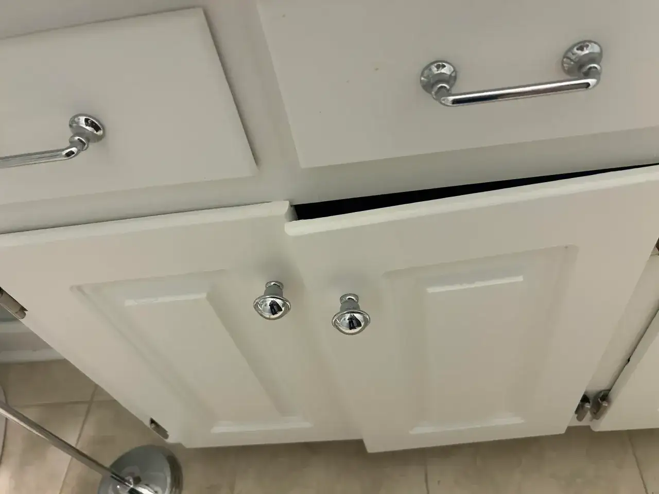 Les portes blanches d'un meuble de salle de bain sont ouvertes, révélant un espace sombre à l'intérieur. Une porte qui ne ferme plus laisse entrevoir le désordre.