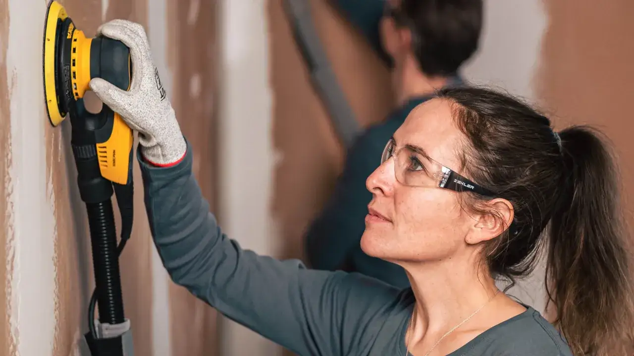 Une femme porte des lunettes et des gants pour poncer un mur avec une ponceuse &eacute;lectrique jaune.