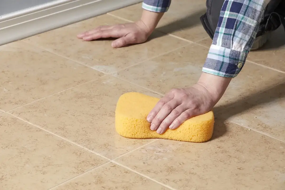 Main tenant utilise une &eacute;ponge jaune pour faire des joints de carrelage sur un sol beige.
