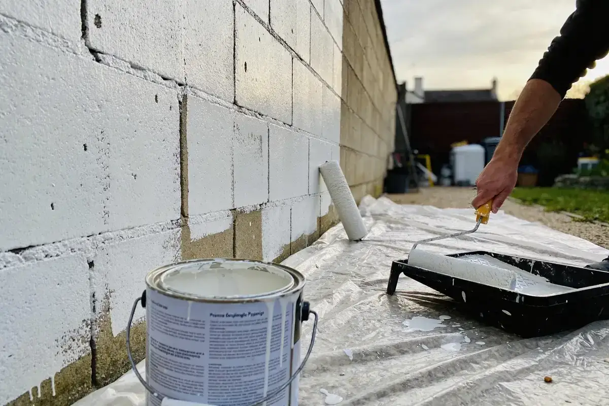 Un homme est en train de peindre un mur en parpaing avec un rouleau. La peinture blanche recouvre progressivement les blocs de b&eacute;ton.