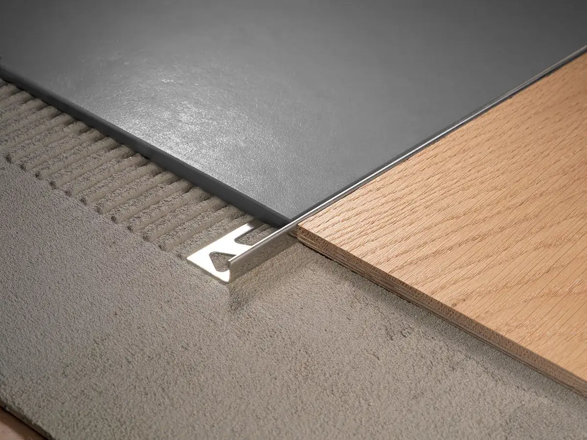 Détail d'une jonction parquet carrelage avec un profilé métallique. Le parquet en bois clair rencontre le carrelage gris posé sur un lit de colle.